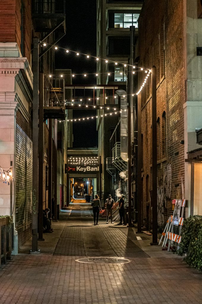 A bustling narrow alley at night featuring string lights, people, and urban architecture.
